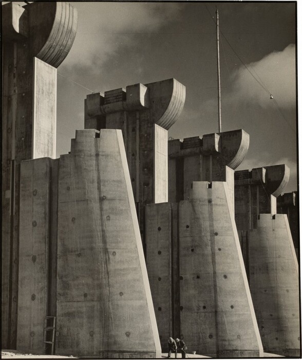 Four monumental concrete structures nearly fill the space of this vertical black and white photograph. The fortress-like architectural forms are arranged in a line so they move away from us at a diagonal to our right to create an almost abstract composition. Each structure seems to be made up of two parts: a form that tapers from a wide base to a narrow top, like a fin. Each of those brace a taller vertical panel, each of which has a semi-circular, crown-like crenellation. The top of the leftmost structure is cropped by the edge of the photograph and its bracing foot reaches halfway across the composition. Two people stand at the base of that structure, barely coming a tenth of the way up the lower bracing structure. Deep shadows on our side of the structures contrast with the brighter areas highlighted by the sun. Two clouds float in the sky above. A tall pole supporting a wire nearly reaches the top edge of the photograph to our right.