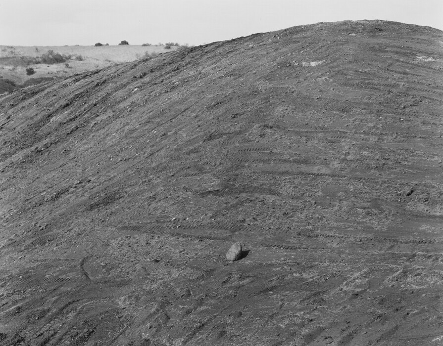 The image shows a gently sloping hill covered with textured earth. The hill dominates the scene, leading the eye upwards. In the distance, sparse vegetation and grass mark the horizon. The terrain is rugged with parallel lines and ridges. The palette is monochromatic, emphasizing different shades of gray. A lone rock is visible in the landscape. The image focuses on the interplay between earth and sky.
