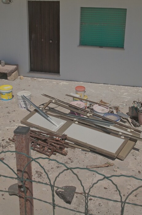The image shows building materials and paint cans in front of a house. Several paint buckets with different colored lids are on a concrete surface in front of a closed, dark wooden door and a window with a green shutter. Nearby, there are metal frames, wooden planks, and a pile of rusty metal objects. A small rock and debris are on the ground, and a green wire fence is visible. The scene conveys a sense of maintenance work or renovation in progress.