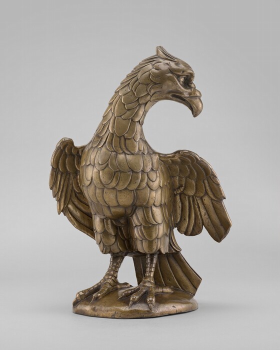 The sculpture depicts a bird with its wings slightly spread and its head turned to the side. It is made of a material resembling bronze, giving it a metallic, golden-brown color. The feathers are detailed, with each layer carefully crafted for a realistic appearance. The bird stands on a textured base, and its eyes and beak are prominent features. The sculpture appears small and designed for indoor display, featuring a shiny surface that reflects light.