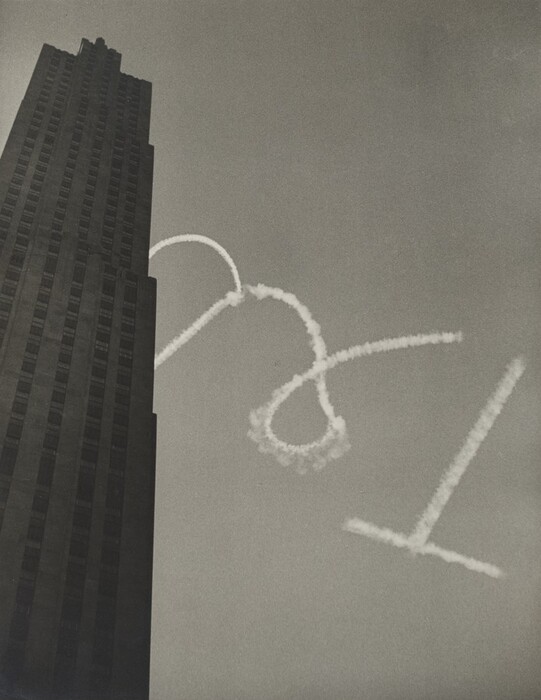 This is a photograph of a tall building with skywriting. The image captures a high-rise building on the left side, its vertical lines extending upwards towards the top edge of the photograph. The clear sky forms the backdrop, while white smoke trails in mid-air create a loop and a curve. The composition contrasts the solid structure of the building with the airy trails of smoke.