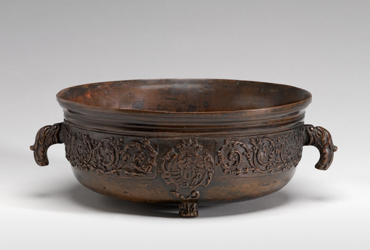 The sculpture is a bronze bowl with two intricately designed handles resembling stylized animal heads, possibly gryphons or dragons. The bowl's surface is adorned with ornate patterns and a central emblem or crest. It is crafted from bronze, giving it a dark metallic hue that highlights its design. The bowl is raised off the ground on three small feet, each matching the decorative scheme.