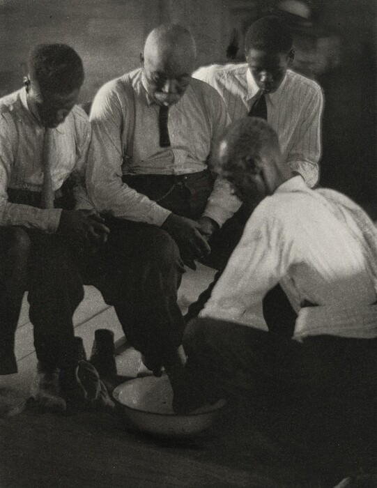 The image shows four men seated or crouching together. The man in the foreground is partially crouching and appears to be performing a task, while the three other men are seated closely next to each other. The man in the middle has a bald head and facial hair, while the men on either side have short, dark hair. All men are wearing long-sleeved shirts with ties; the shirts have light, possibly striped patterns. There is a bowl on the floor, and the man who is crouching is using it for some activity involving his hands. The environment appears to be indoors, with a dimly lit background that suggests a simple, perhaps wooden interior.