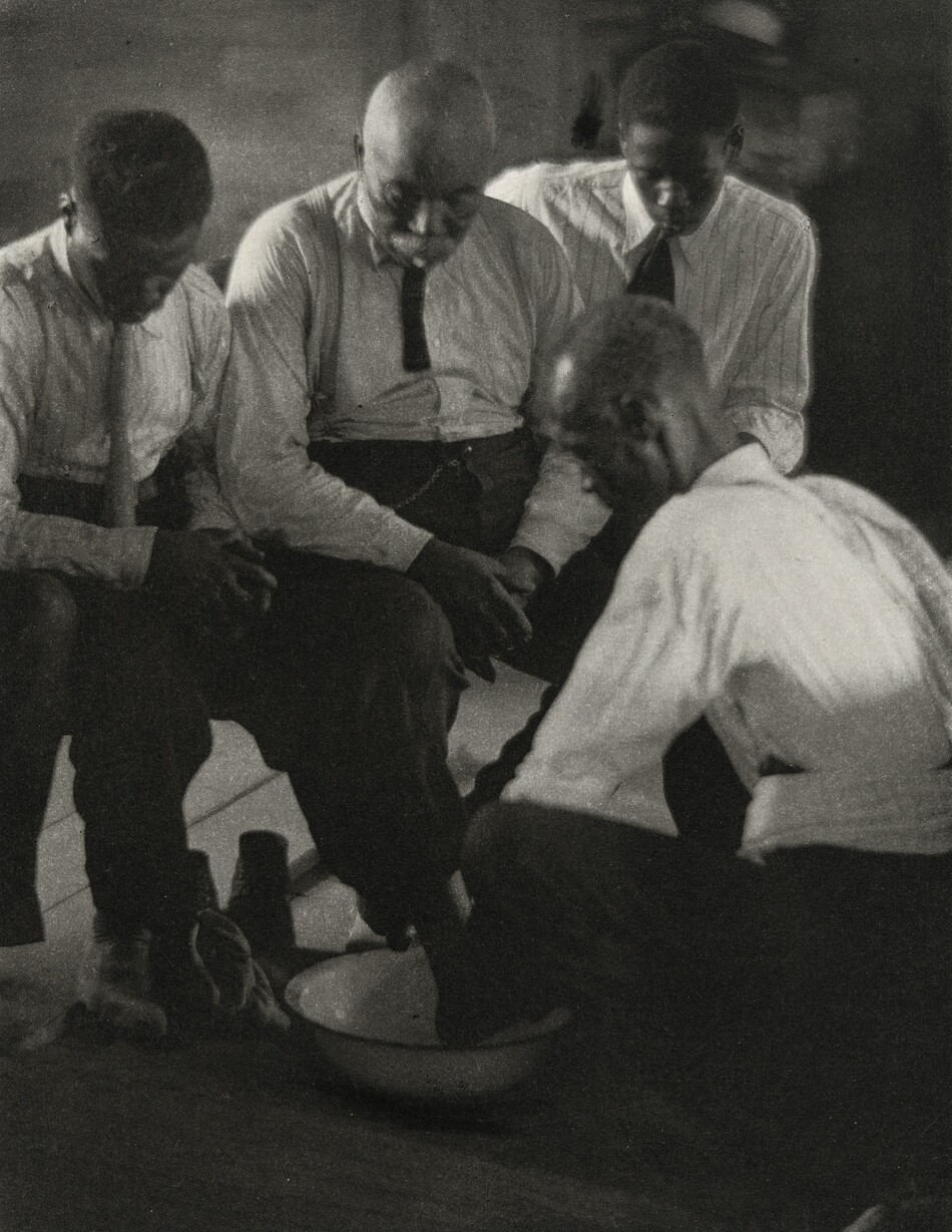 The image shows four men seated or crouching together. The man in the foreground is partially crouching and appears to be performing a task, while the three other men are seated closely next to each other. The man in the middle has a bald head and facial hair, while the men on either side have short, dark hair. All men are wearing long-sleeved shirts with ties; the shirts have light, possibly striped patterns. There is a bowl on the floor, and the man who is crouching is using it for some activity involving his hands. The environment appears to be indoors, with a dimly lit background that suggests a simple, perhaps wooden interior.