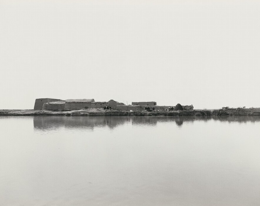 This image shows a settlement across a calm body of water. The horizon is high in the composition, highlighting the reflective water in the foreground. Various buildings and structures are visible along the horizon line. The scene is captured photographically, with crisp detail and clear lines. The color palette is monochromatic, with shades of gray creating a tranquil mood. The sky merges with the land in the distance.