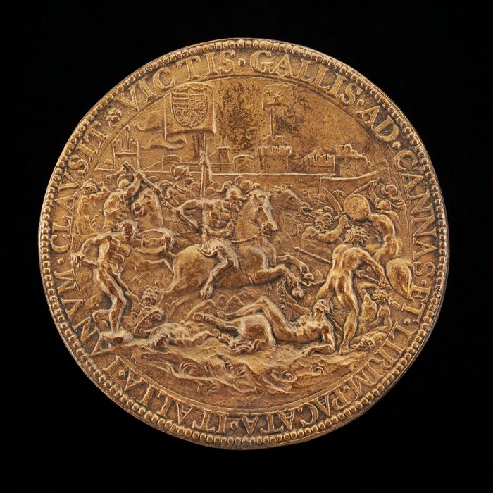 The sculpture features a circular medal with a detailed scene of battle. The scene includes soldiers on horseback and infantry engaging in combat. The medal is golden-hued, giving it a metallic appearance. Details include figures in action on horses and foot, some appearing minimally clothed. In the background, faint structures hint at a battlefield setting. Inscribed text along the edge adds to its historical or commemorative significance.