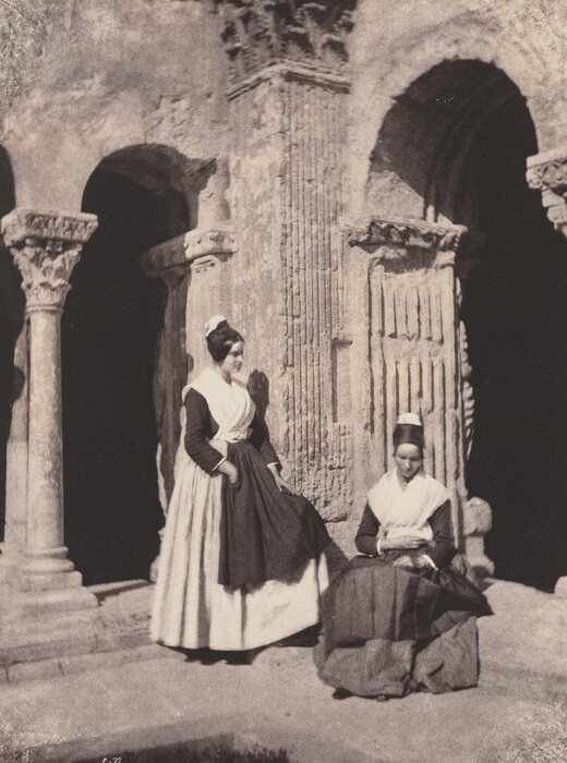 The image shows two women in full-length, dressed in period clothing. One woman is standing, leaning against a carved stone structure with her one arm holding up a portion of her long dark dress. Her other arm is bent, resting on her hips. She wears a white bonnet, white apron, and a scarf-like accessory around her shoulders. The other woman is seated, turned slightly to the side with her hands folded in her lap. She wears a dark dress, white apron, and a bonnet. The background consists of architectural elements that appear ancient or in a classical design, featuring columns and arches, with intricate carvings and textures on the stone. The scene is set in an exterior location, possibly an ancient ruin, with shadows suggesting an afternoon light.