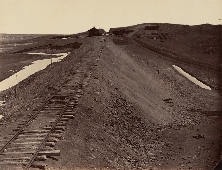 The image shows a railway track stretching into the distance. The track is positioned towards the left edge of the image, running diagonally towards the horizon, which is placed around the upper third of the image. A small building sits on the horizon line, near the center of the image. The terrain on either side of the railway appears rocky and barren, with a few small pools of water visible to the left. The color palette is predominantly sepia tones, giving the image a vintage appearance.
