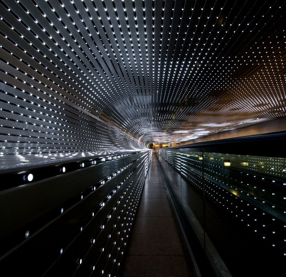 We look through a tunnel lined with dozens of strings of bright white lights nestled in silver-gray slats that zoom away from us in this horizontal photograph. At the far end of the tunnel, at the center of the photograph, the alternating lines of lights and slats almost come together around a narrow, capsule-shaped area of golden yellow light. The flat ceiling curves down to meet the wall to our left. The curve continues to make a C-shape before extending straight down the remaining height of the wall, turning the profile of the open space into a backward-facing P. Some of the lights are bright, some fading, and some are off to create a loose pattern of dark squares floating in a field of light. The lights seem brighter at the far end of the tunnel. The overall impact of the view is a starburst radiating in diagonal lines, coming from a point at the center of the photograph.