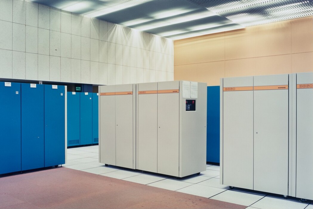This is a photograph of a computer server room with data cabinets. The room is well-lit and filled with large server cabinets. The cabinets are white and blue, some labeled with the brand "Siemens." The floor is covered with white tiles and some areas have a brownish carpet. The ceiling has linear lighting fixtures casting a muted light. The walls are lightly textured with beige and white tones.