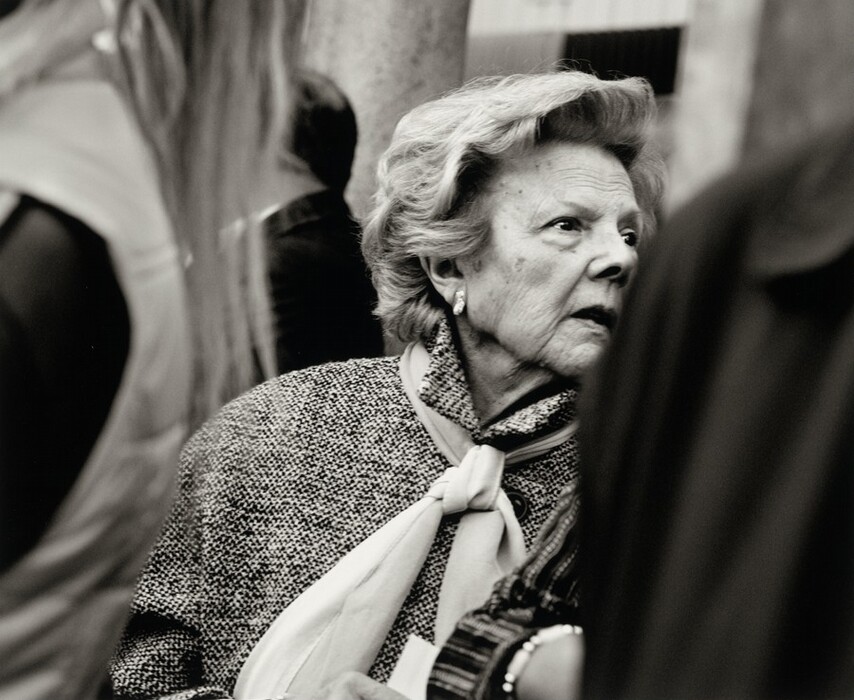 The image depicts an elderly woman in a half-body view. She is in an upright position, facing slightly to the right, with her head turned towards the camera. She has visible wrinkles, a small nose, and short, wavy, light-colored hair. The woman is wearing a textured coat and a tightly tied scarf around her neck. There is a partial view of a bracelet on her wrist. People are on either side of her in the crowded area, and there is a blurred exterior environment in the background, suggesting the image was taken outdoors.