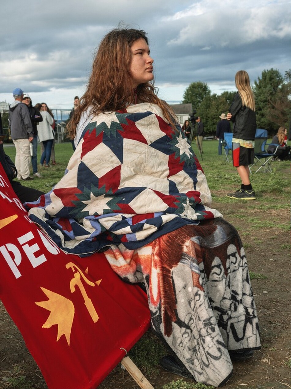 This photograph shows a person sitting outdoors, their torso wrapped in a quilt with a geometric pattern in red, white, and blue with star designs, and their legs wrapped in an additional blanket featuring Star Wars characters. The person's face is in profile, looking to the right. They have pale skin and long, wavy brown hair. They are seated on an unseen surface, likely a chair, on top of the grass, and there's a red banner with yellow symbols and white text partially visible beside them on the far left. In the background, several other people are standing or sitting, with trees and a cloudy sky overhead.