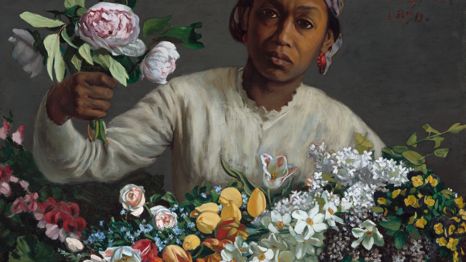 Holding a bunch of peonies in one hand, a woman with brown skin leans forward, toward us from behind a large basket holding dozens of flowers in this horizontal painting. The basket holding the flowers spans the width of the canvas, and the woman is shown behind it from the chest up. She wears a cream-white, long-sleeved blouse with scalloped trim around the high neck. She wears coral-red earrings, and a plaid cloth in tones of rust red, slate blue, pale purple, and black is tied tightly over her black hair, which is visible over her ears. Her brow is slightly furrowed, and she looks at us with large, dark eyes. Her full mouth is closed, the corners faintly downturned. She reaches her right arm, on our left, toward us with a bouquet of three pink-and-white peonies and greenery. Her basket is filled with yellow and red tulips, pink roses, white and purple lilac, and other white, pink, yellow, and blue flowers, and it takes up the bottom third of the composition. The woman and basket are shown against a dove-gray background. The artist prominently signed and dated the work with red letters near the upper right corner, near the woman’s head: “F. Bazille. 1870.”
