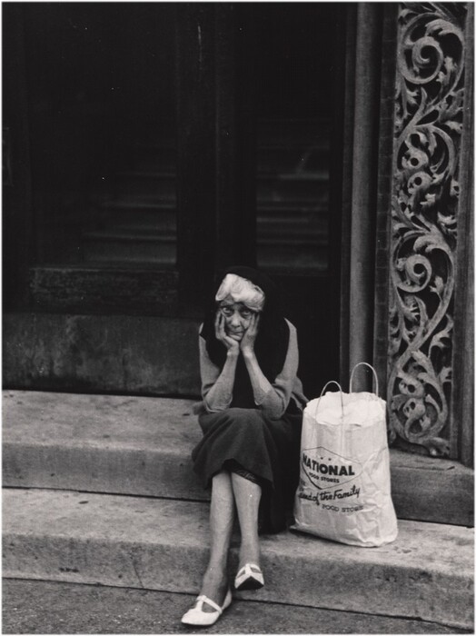 The image shows an elderly woman seated on a concrete step. She is leaning forward with her elbows resting on her knees and her hands supporting her face. The woman's facial features are discernible; she has a lined face and is wearing a headscarf that covers most of her hair. Her outfit consists of a long dress or coat, dark-colored hosiery, and light-colored flat shoes. Next to her on the step is a large paper shopping bag. The background features a decorative building facade with intricate carvings to her right and a large, dark doorway behind her.