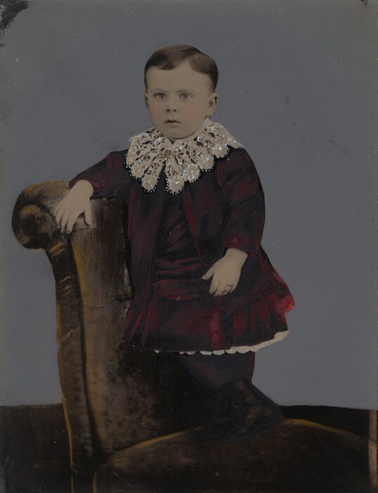 The image shows a child posing while standing on an upholstered chair. The child has short, neatly combed brown hair and features a calm expression. They are dressed in a dark, long-sleeved garment with a large lace collar, dark stockings, and shoes. The chair is upholstered in dark fabric and the background is a neutral gray.