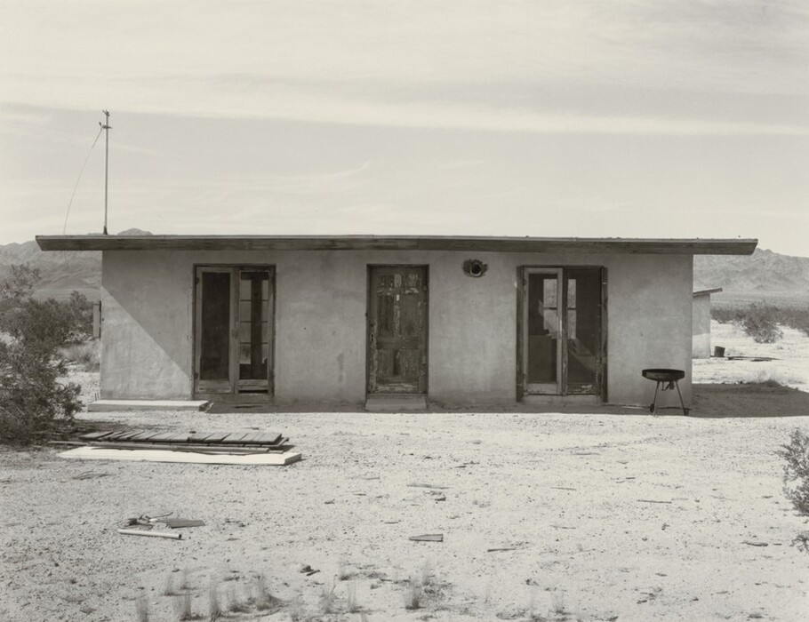 The image shows a frontal view of a single-story building. The horizon is positioned above the roofline of the building, with distant mountains in the background. The building has a flat roof, two doorways, one window, signs of wear on the exterior, an antenna on the left and a small barbeque grill on the right. The style is photographic, with a monochromatic palette of soft grays. The landscape beyond the building is barren and rocky, with scattered shrubbery.