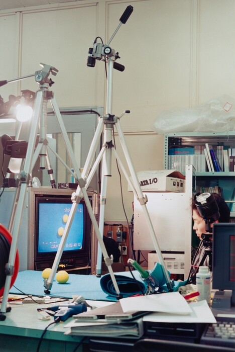 This is a photograph of a technical laboratory setup. The image shows a room filled with various equipment typically found in a scientific or media lab. Two tripods stand in the foreground, one supporting a camera, capturing an experimental setup on a table covered with a bright blue surface. On the table are objects, including tennis balls, also displayed on a nearby screen. The screen shows a recorded view of these objects with a blue background. To the right are technical paraphernalia, including wires, notebooks, and electronic devices like a light fixture. In the background are shelves with binders and boxes. The setting portrays a controlled environment for recording or testing, emphasizing the technical aspect of the laboratory.