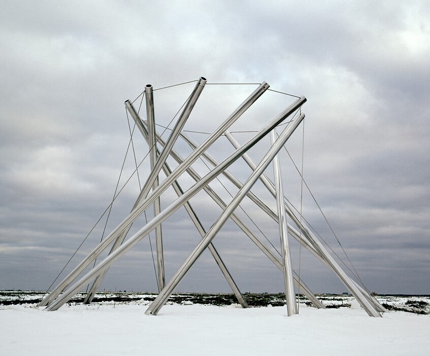 This sculpture is made up of ten long, stainless steel tubes standing upright in a ring. Each upright tube angles to the right and is tethered by one wire cable that connects the top edge to the ground and another that connects with the top of the next tube, creating a ring around the top edges. The tubes start to converge like a shutter but are stationary. In this photograph they stand on a snowy field under a cloud-carpeted sky.