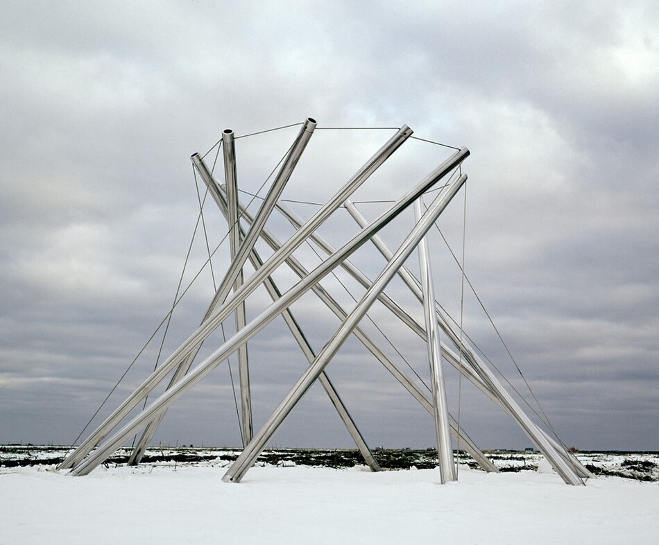 This sculpture is made up of ten long, stainless steel tubes standing upright in a ring. Each upright tube angles to the right and is tethered by one wire cable that connects the top edge to the ground and another that connects with the top of the next tube, creating a ring around the top edges. The tubes start to converge like a shutter but are stationary. In this photograph they stand on a snowy field under a cloud-carpeted sky.