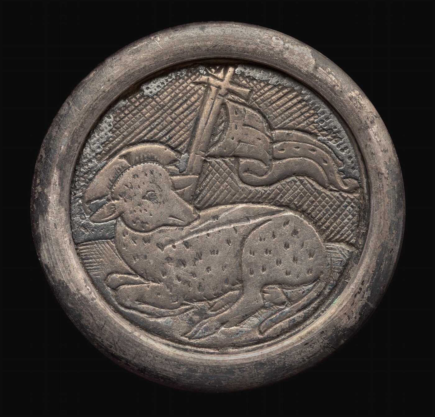 The sculpture portrays a relief of a lamb with folded legs and a curled horn. Behind the lamb is a flag with a cross on a pole, all encircled by a round frame. The sculpture is made of a metallic material with a silver/pewter color, showing a weathered texture with tarnishing and signs of wear. The background has a textured pattern contrasting with the smoother depiction of the lamb and flag. The design is somewhat flat, with the subjects in low relief, making it more decorative than three-dimensional.