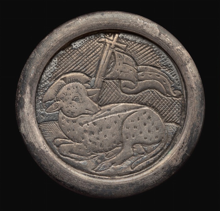 The sculpture portrays a relief of a lamb with folded legs and a curled horn. Behind the lamb is a flag with a cross on a pole, all encircled by a round frame. The sculpture is made of a metallic material with a silver/pewter color, showing a weathered texture with tarnishing and signs of wear. The background has a textured pattern contrasting with the smoother depiction of the lamb and flag. The design is somewhat flat, with the subjects in low relief, making it more decorative than three-dimensional.