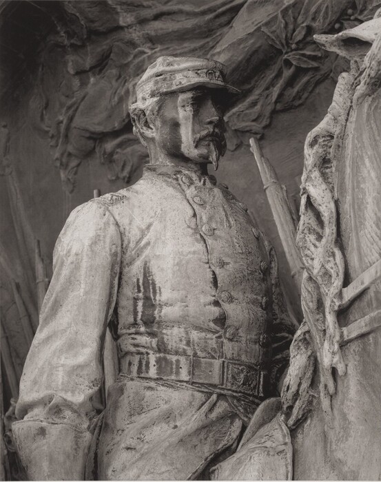 The sculpture depicts a figure in a military uniform standing alongside a horse. The attire is detailed, including a coat, cap, belt, and hat. The figure stands upright in a dignified pose. The sculpture is primarily monochromatic and appears weathered. The background seems to be part of a relief. The portrayal is realistic in terms of human proportions and expressions.
