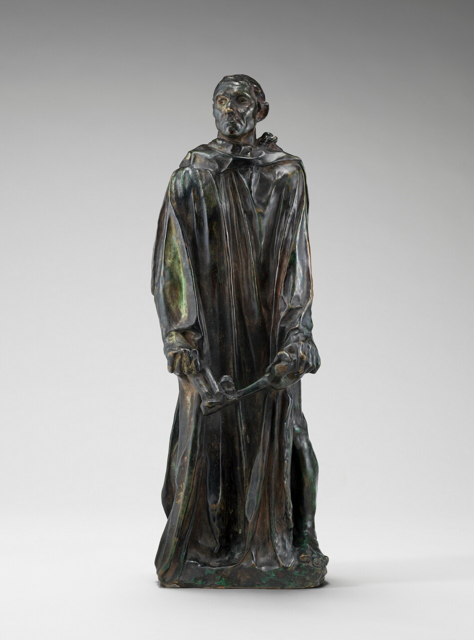 Cast in bronze, this free-standing sculpture shows a man with a gaunt face wearing a robe that falls in long, vertical folds as he holds an oversized skeleton key down in front of his thighs. In this photograph, the man’s body faces us but he turns his head to our left to look in that direction, his chin sightly raised. He has close-cropped hair, sunken eyes, hollow cheeks, a wide, square chin, and his thin lips are downturned. The long-sleeved robe drops heavily to his feet, and the toes of one bare foot peek out. A hint of a raised, round halter like a yoke around his neck shows from under his robe near his left shoulder, on our right. Most of the surface has a dark green patina except for a few lighter places, as on his sleeve to our left and in the thick drapes of his robe. A few areas are rubbed gold, including the tip of his nose and the knuckles of his hands.