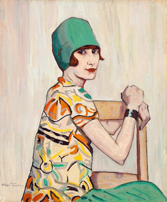 This painting depicts a woman from the waist up, seated sideways on a wooden chair. She is turned to the right, with her hands on the back of the chair, and looks over her shoulder towards us. She has pale skin and short reddish-brown hair visible beneath a green cap. Her red lips are slightly turned up at the edges, and her eyes are dark. She is wearing a brightly colored top with abstract shapes in orange, yellow, gray, and green against a cream background, and a green skirt that is the same shade as her hat. A bracelet adorns her wrist with vertical stripes of white, dark blue, and dark red. The chair she sits on is light yellow wood, and the background is made of vertical lines of white, cream, and gray, with hints of pastel pink, minty green, and pale blue. The brushstrokes in the painting are visible.