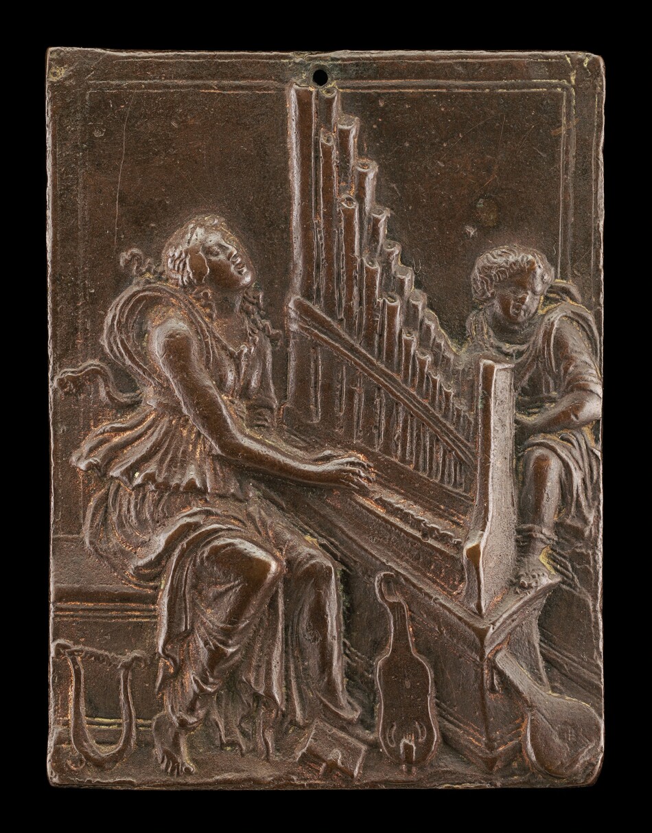 The sculpture is a relief depicting two figures seated and playing an organ. The figure on the left is a woman, dressed in flowing garments, playing the organ with hands positioned on the keyboard. The figure on the right is behind the organ. The sculpture is bronze in color, suggesting it may be made of bronze. The surface has a patina typical of metal sculptures. The attention to detail in the attire and instrument gives the scene a sense of activity and interaction around music. Additional elements include a lyre and another stringed instrument at the base. The organ pipes are arranged vertically at the center, emphasizing the musical theme.
