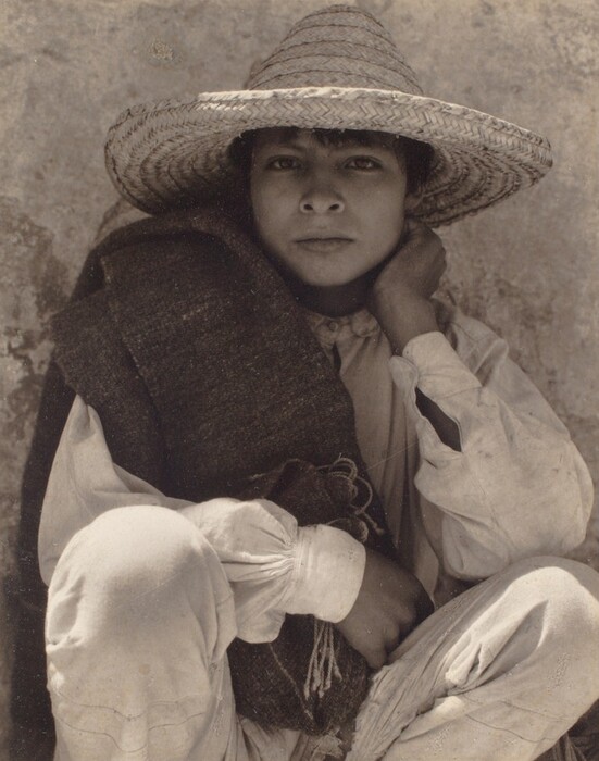 The image shows most of the body of a person sitting with one knee drawn up, facing forward. Their right hand is resting against their cheek. The individual has short hair under a wide-brimmed, woven hat. They wear a loose-fitting, long-sleeved shirt and matching pants made of light-colored fabric. Draped over their shoulder is a dark-colored woven garment or fabric. The background is an aged, textured surface resembling a stone or concrete wall.