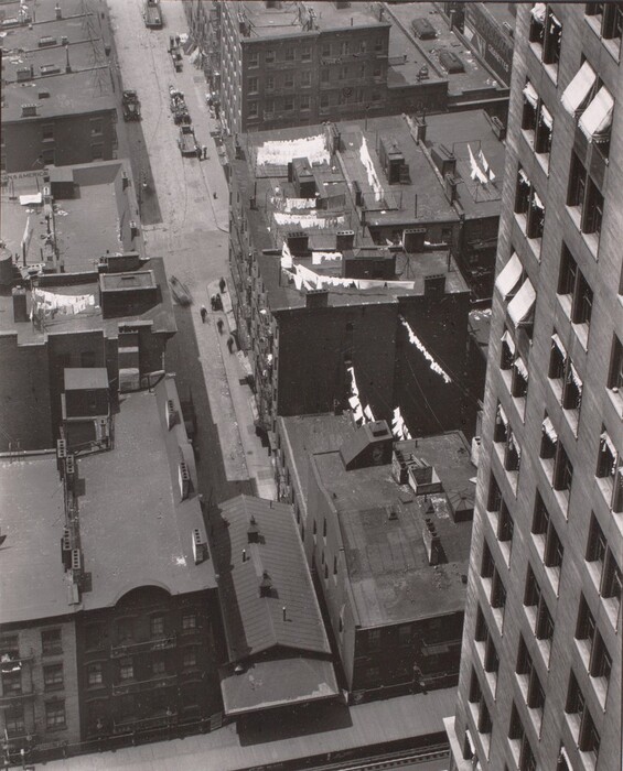 This is a photograph of an urban street scene viewed from above. The image captures a city block with uniform, flat-roofed buildings tightly packed together. In the foreground, there's a rooftop with an irregular, chimney-covered surface. Laundry hangs on visible lines strung along the rooftops. The street is lined with several multi-story buildings, each with rows of windows reflecting light and shadows. The streets below are populated with vehicles, likely cars or streetcars, with two separate streets visible, running parallel and intersecting at the base of the image.