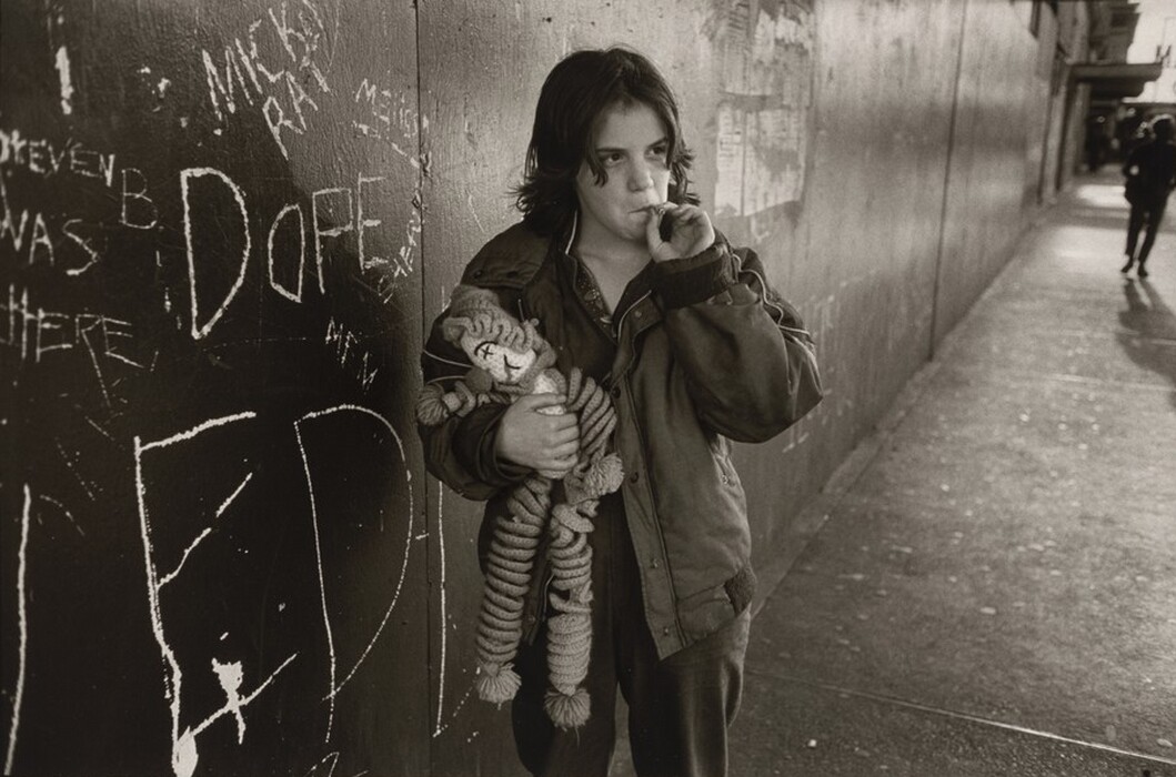 The image shows a person standing against a wall. The person has medium-length dark hair and is smoking while looking off to the side. They are wearing a dark jacket over a shirt and holding a plush toy with a large smile embroidered. The wall beside them is covered in chalk graffiti, with various words and names visible. In the background, a sidewalk stretches back into the distance, with another person walking away.