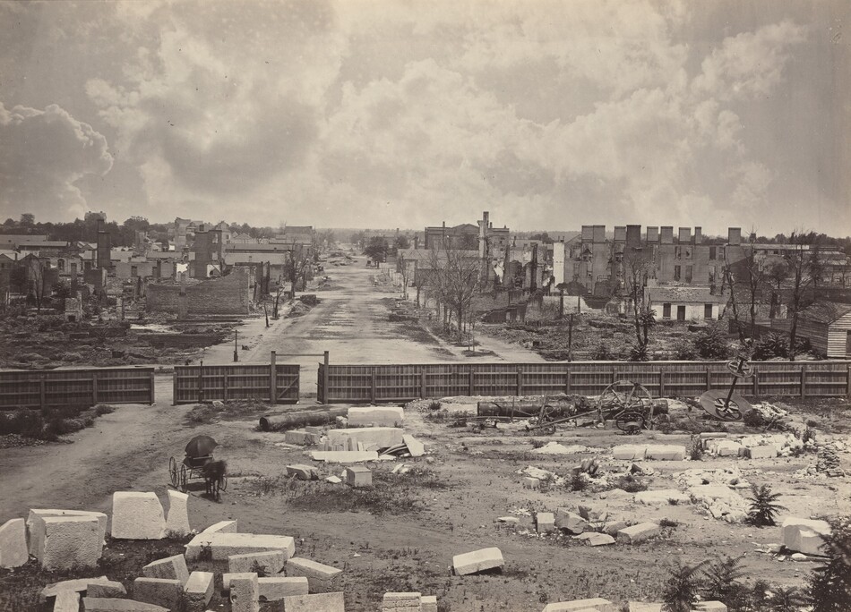The image shows an aerial view of a landscape with ruins of a city. There are scattered large stone blocks, remnants of machinery, and a wooden fence marking a boundary. A deserted street leads into the distance, surrounded by charred and skeletal remains of buildings. Trees and faint structures can be seen on the horizon. The sky is filled with large clouds, casting somber light over the scene. The photograph's monochromatic palette emphasizes the contrast between sky and ruins.