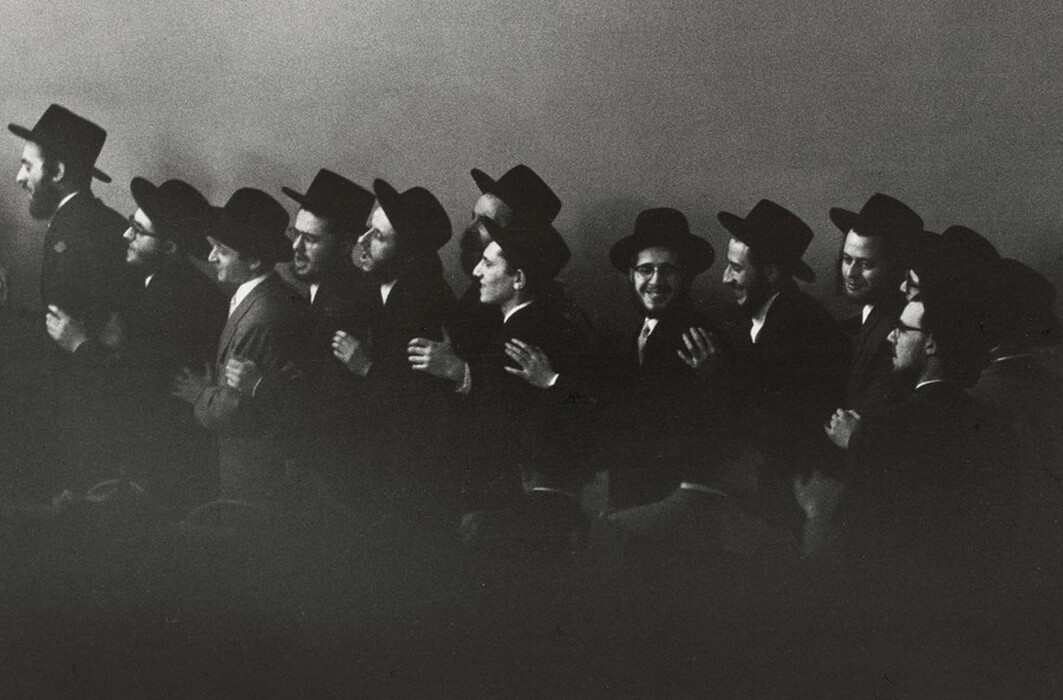 The image shows a group of men in a medium shot, as their upper bodies are visible. They are positioned in a line, closely standing shoulder to shoulder, some with their arms linked or hands placed on each other's shoulders. The men have distinct facial features and are wearing traditional hats and formal attire, including suits and ties. Their hair is mostly hidden under their hats. The background is plain, suggesting an indoor setting without distinct decorative elements. No specific jewelry is visible, and the lighting casts soft shadows, enhancing the group's cohesion.