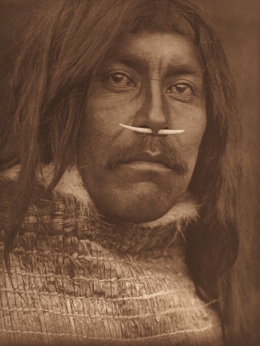 The image is a close-up portrait of a person's face and shoulders. The individual faces forward with prominent cheekbones, a strong jawline, and eyes looking into the camera. They have long, textured hair and wear a horizontal nose ornament. The person's clothing is textured, and the background is neutral.