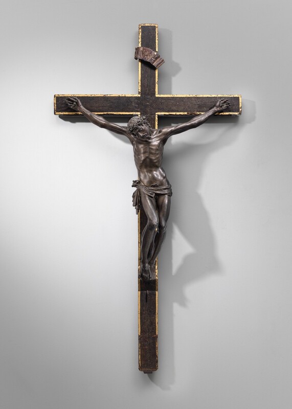 The body of a man cast from bronze hangs heavily from nails driven through his hands and both feet on a wooden cross in this sculpture, which would hang on a wall. The man’s head hangs down to our left, eyes closed as his shoulders shift to our left and his hips to our right. A ring of thorns encircles long, wavy hair. His torso and legs are lean and muscular, and he wears only a loincloth tied across his hips. His arms are unnaturally long and sinewy. A bronze banner with the letters “INRI” is affixed to the vertical beam near the top of the cross. The surface of the wood cross is dinged and speckled with worm holes, and the edges of the face of the cross are painted gold.