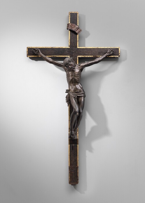 The body of a man cast from bronze hangs heavily from nails driven through his hands and both feet on a wooden cross in this sculpture, which would hang on a wall. The man’s head hangs down to our left, eyes closed as his shoulders shift to our left and his hips to our right. A ring of thorns encircles long, wavy hair. His torso and legs are lean and muscular, and he wears only a loincloth tied across his hips. His arms are unnaturally long and sinewy. A bronze banner with the letters “INRI” is affixed to the vertical beam near the top of the cross. The surface of the wood cross is dinged and speckled with worm holes, and the edges of the face of the cross are painted gold.