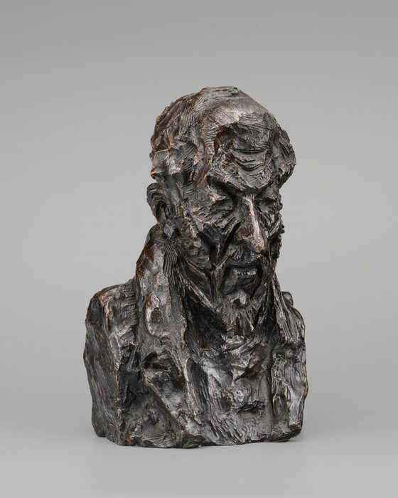 The sculpture is a bust of a man with a beard and wrinkled skin. The man has a long nose, deep-set eyes, and thinning hair. He appears to be wearing a jacket with a high collar. The sculpture has a rough surface, with lots of visible marks.