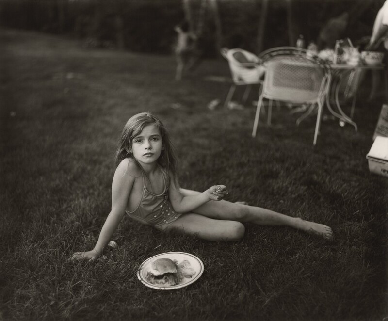 A young girl half lies, half sits in grass in this horizontal, black and white photograph. The girl wears a one-piece bathing suit and sits propped on her right arm, to our left. One leg stretches long to the right and the bottom knee is bent. She looks directly at us with dark eyes, and her lips are closed. Her long hair falls behind her shoulders. The other elbow angles into her belly, and she holds something small, perhaps a potato chip, in that hand out to the side. A plate with a hamburger bun and chips sits in the grass in front of her. Chairs and a table laden with dishes and bottles are out of focus in the background, in the top right quadrant of the photograph, and a child beginning to climb a tree is barely discernable beyond that.