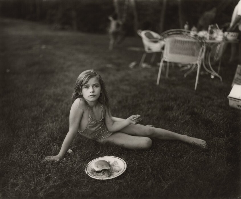 A young girl half lies, half sits in grass in this horizontal, black and white photograph. The girl wears a one-piece bathing suit and sits propped on her right arm, to our left. One leg stretches long to the right and the bottom knee is bent. She looks directly at us with dark eyes, and her lips are closed. Her long hair falls behind her shoulders. The other elbow angles into her belly, and she holds something small, perhaps a potato chip, in that hand out to the side. A plate with a hamburger bun and chips sits in the grass in front of her. Chairs and a table laden with dishes and bottles are out of focus in the background, in the top right quadrant of the photograph, and a child beginning to climb a tree is barely discernable beyond that.