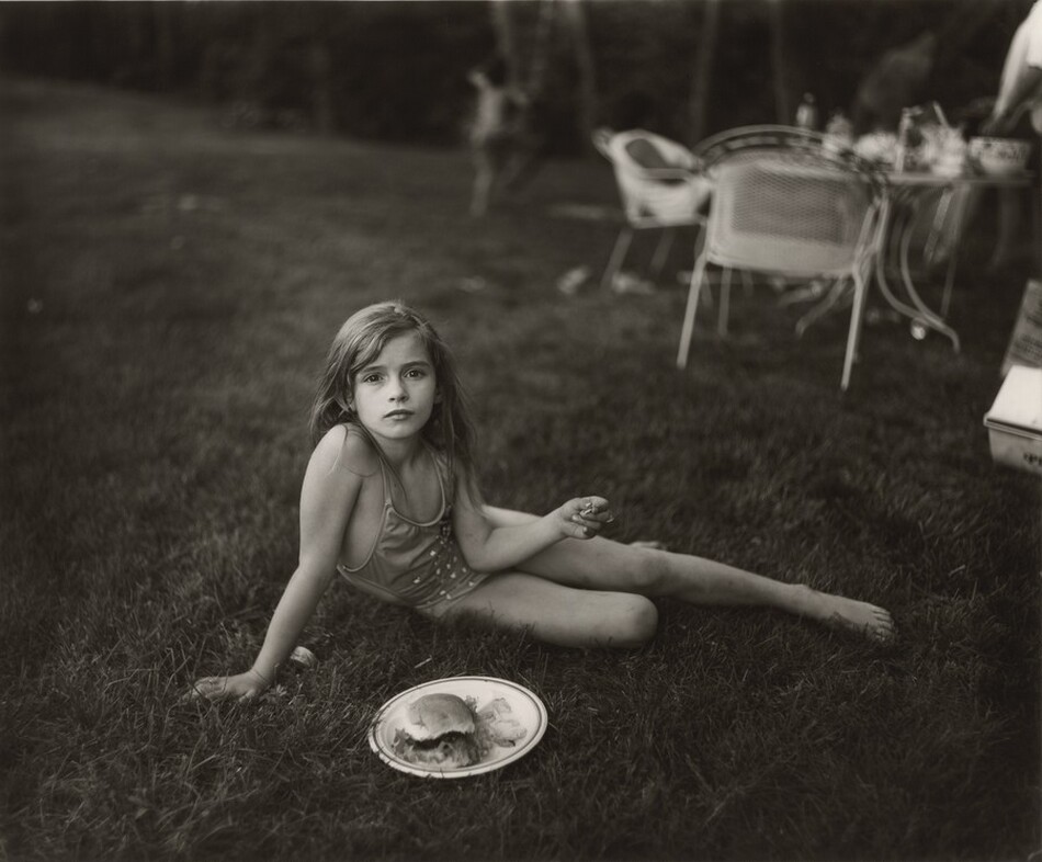 A young girl half lies, half sits in grass in this horizontal, black and white photograph. The girl wears a one-piece bathing suit and sits propped on her right arm, to our left. One leg stretches long to the right and the bottom knee is bent. She looks directly at us with dark eyes, and her lips are closed. Her long hair falls behind her shoulders. The other elbow angles into her belly, and she holds something small, perhaps a potato chip, in that hand out to the side. A plate with a hamburger bun and chips sits in the grass in front of her. Chairs and a table laden with dishes and bottles are out of focus in the background, in the top right quadrant of the photograph, and a child beginning to climb a tree is barely discernable beyond that.