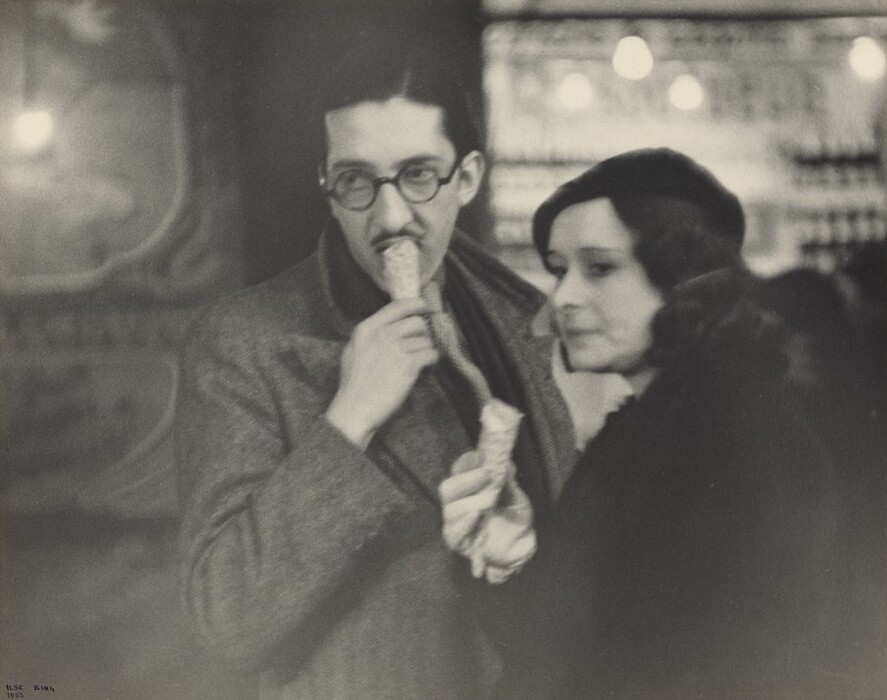 The image shows two individuals standing next to each other from the waist up. The man on the left holds an ice cream cone near his mouth, while the woman on the right leans slightly towards him. The man has short, dark hair and wears round glasses and a dark, patterned overcoat. The woman has dark wavy hair, and she wears a dark coat with likely fur trim and a hat. The background is blurred with soft lighting. Neither individual is wearing jewelry.