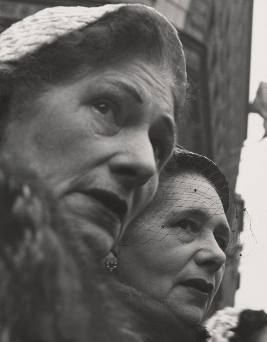 The image depicts a close-up of the upper bodies of two women, focusing on their faces. Both women are closely positioned, with the woman on the left slightly in the foreground. The woman in the front is wearing a textured hat and has prominent features, including defined eyebrows and lips set in a serious expression. The woman behind has her hair neatly styled back and is adorned with a netted veil over her face, with a similarly serious expression. Both women appear to be wearing fur coats, with the textures visible in the foreground. The woman on the left is wearing small, bead-like earrings. The background shows an indistinct view of a building and a flag.