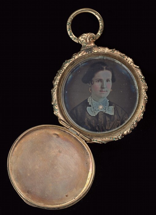 The image depicts a portrait of a person in a small locket. The portrait shows the person from the shoulders up, facing the viewer with a slight angle. The individual has fair skin, dark hair styled neatly around the head, and is wearing a dark garment with a white lace collar. The person has gold earrings and a jeweled brooch at the neck. The portrait is encased in an ornate round metal frame with a floral or leaf design, with a hinged metal cover opened to reveal the portrait. The background of the portrait is plain, and the locket is set against a black background.