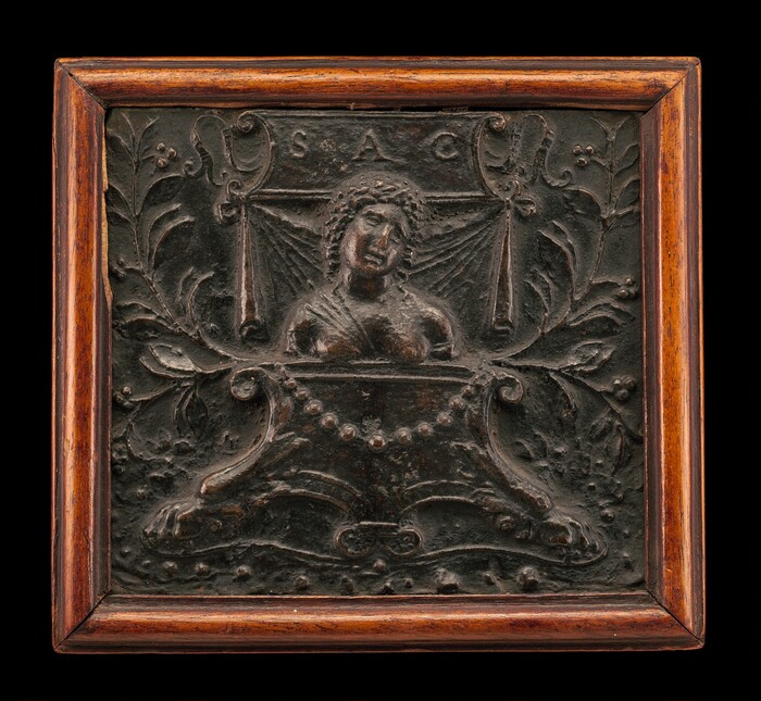 The sculpture depicts a carved relief showing the upper body of a figure with curly hair emerging from an ornate structure resembling a podium or urn, adorned with beads. Intricate leaf and scroll motifs decorate the background, with draped fabric elements on each side. Inscribed at the top are the letters 'SAC'. Crafted from a dark material, possibly bronze or iron, it is set in a square wooden frame with a reddish-brown hue. The relief combines classical and decorative elements, featuring detailed textures and intricate designs.