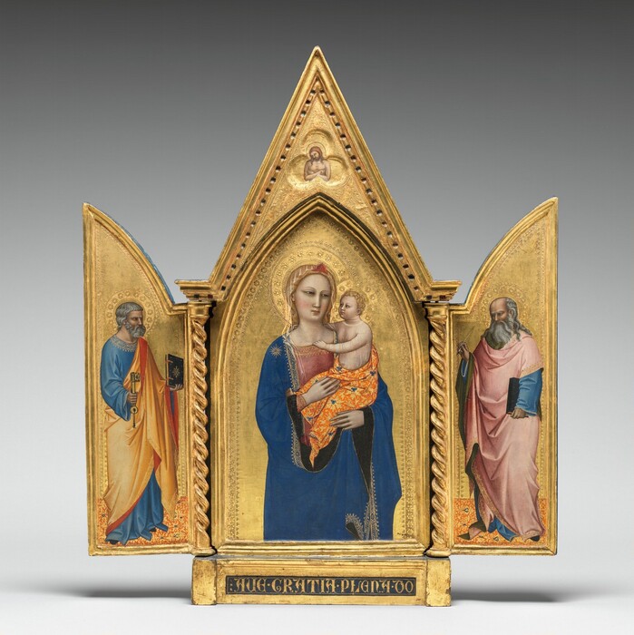 Two panels on a gilded, carved, wooden frame are swung open like shutters to show a bearded, gray-haired man wearing robes standing in each wing, on either side of panel with a pointed, arched top showing a woman holding a baby. In a three-lobed shape in the apex above the woman and child, a nude man crosses his arms over his chest. All the people have pale, ivory-colored skin. Gold halos encircle each person’s head and the background of all three panels is bright gold. The woman in the center panel is shown from the hips up with her body facing us. The deep blue mantle that drapes over her shoulders and her dusty rose-pink dress beneath are trimmed in gold. A sheer veil covers her wavy blond hair and a red, triangular form sits above her forehead like a diadem. She looks to our right with dark, almond-shaped eyes. She has a straight nose, her coral-pink lips are closed, and her smooth cheeks are lightly flushed with pale pink. She holds the child in the crook of her left arm, on our right, and rests her other hand in the baby’s lap. He has short, curly blond hair and looks directly at the woman. He reaches his right arm, farther from us, around his mother’s neck and hooks his other hand in the neckline of her dress. He wears a sheer shirt edged in gold, and the fabric draped around his hips is patterned with tangerine-orange and gold, with lapis-blue clover designs. He has rounded cheeks and a pudgy tummy, but his facial features and arms are scaled more like an adult. Twisted columns separate this central panel from the shutter-like panels to each side. A single person stands in each wing, filling most of the space, and both are angled and look towards the central panel. Both men have darker, olive-toned complexions, wear long robes trimmed in gold, and stand on floors patterned with orange, gold, and sparsely spaced royal blue diamond-shapes. The man to our left has short, steel-gray hair and a trimmed beard. Wrinkles line his forehead, and his brow is furrowed. He wears a topaz-blue, long sleeved garment under a honey-orange robe, the underside of which is ruby-red where it turns back over his wrists and shoulder. He holds a book with a dark cover up in his left hand, farther from us, and a large skeleton key in his other hand. In the right panel, a balding man has a fringe of long, gray hair and a flowing, full beard. His forehead is also creased with wrinkles and his brow is furrowed. The dark, olive-green lining of his rose-pink robe shows where it folds back at his neck, over his wrists, and down the front. His robe covers a topaz-blue garment as well. He holds a book, also with a dark cover, by his side with his left hand, closer to us, and holds up a feather quill with his opposite hand. The inner edges of each of the central panel and both wings are incised with bands of decorative floral and geometric designs punched into the gold backgrounds. In the pointed peak above the central panel, the nude man is shown from the waist up in a three-lobed, clover shape that would be visible even if the wings were closed. The man’s body faces us but his haloed head tips to his right, our left. He has a beard and long brown hair, and a hole dripping blood pierces the back of his left hand, visible on our right. The base of the tryptic is inscribed with gold letters in a dark field: “AVE GRATIA PLENA DO.”