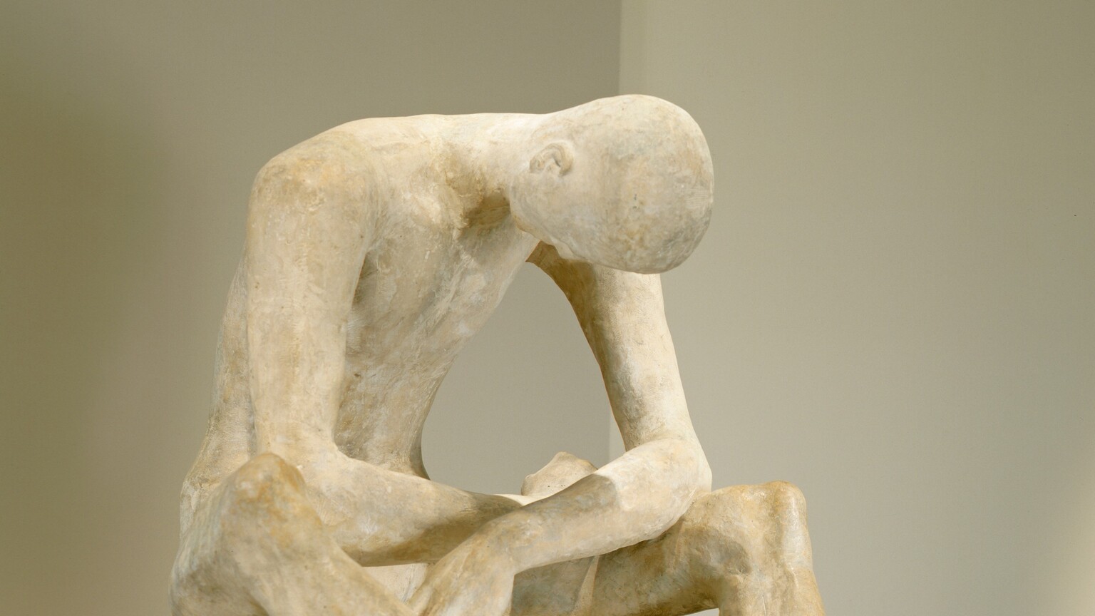 A man bows his bald head deeply as he rests his splayed elbows on his thighs in this freestanding plaster sculpture. The long toes of the man’s feet nearly touch, and they are angled to our right in this photograph. He sits on a low surface, also sculpted from plaster, and his long limbs create a cage around his hollowed chest. The surface of the plaster is ivory white in some areas and parchment brown in others. The piece rests on a gray pedestal against gray walls.
