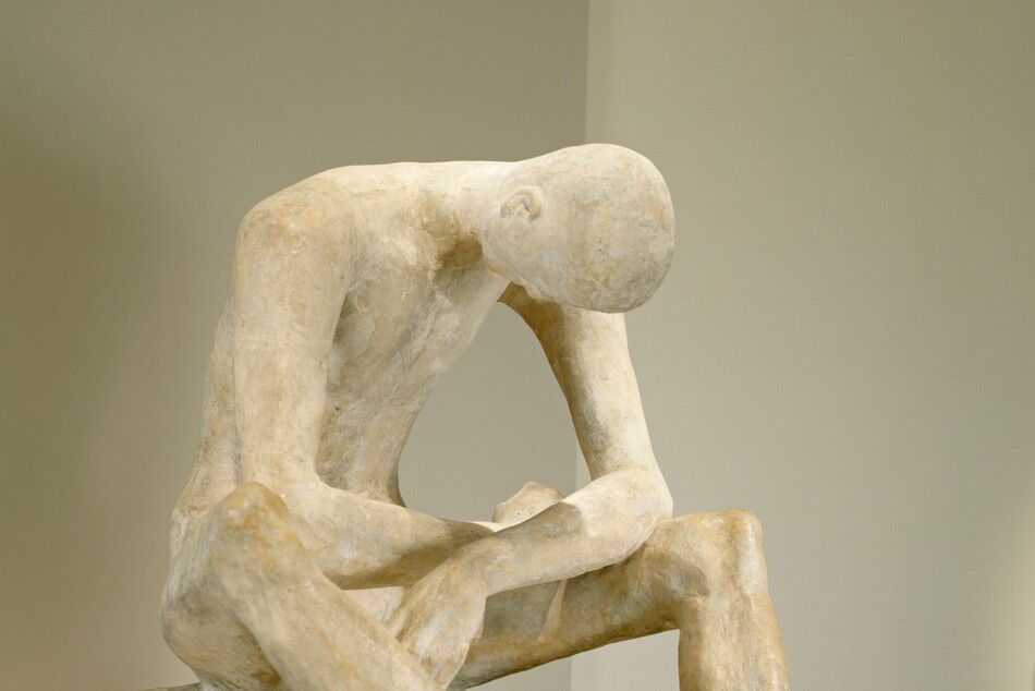 A man bows his bald head deeply as he rests his splayed elbows on his thighs in this freestanding plaster sculpture. The long toes of the man’s feet nearly touch, and they are angled to our right in this photograph. He sits on a low surface, also sculpted from plaster, and his long limbs create a cage around his hollowed chest. The surface of the plaster is ivory white in some areas and parchment brown in others. The piece rests on a gray pedestal against gray walls.