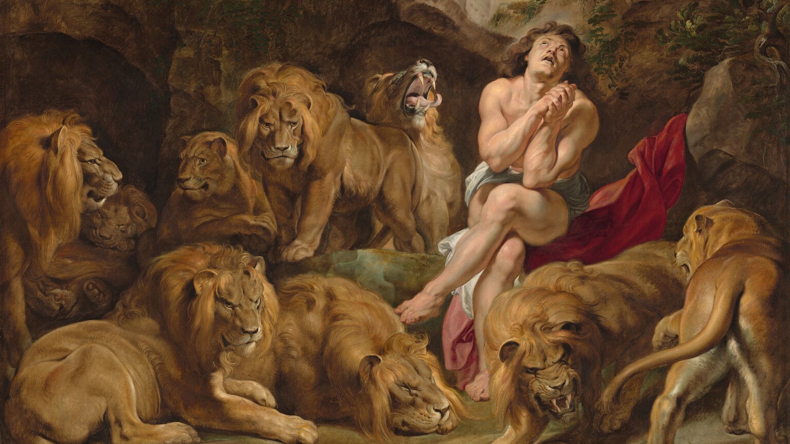 A nearly nude, muscular man with pale, peachy skin sits in an underground cave among seven male and two female lions in this horizontal painting. The man sits to our right of center with his legs crossed, elbows close to his body with hands clasped by his chest. His head tips back to look up through a small opening above. He has long, wavy, chestnut-brown hair and dark eyes. A white cloth wraps across his hips, and he sits on a scarlet-red swath of fabric draped up over a rock next to him. The nine lions stalk, sit, or lie down around the man. One male lion next to the man, to our left, opens his mouth with his head thrown back, curling tongue extended beyond long fangs. A human skull and other bones are strewn on the dirt ground close to us. The rocky cave curves up around the animals and man to a narrow, round opening showing blue sky above.