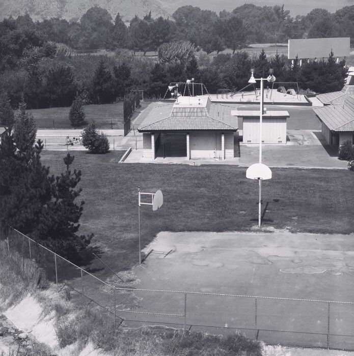 The image depicts an outdoor recreational area from an elevated point of view. The horizon is in the upper third of the image, partly concealed by rows of trees, with hills in the background. A fenced-off basketball court with a white hoop appears in the foreground, surrounded by grassy areas and a couple of small buildings with tiled roofs. Beyond these structures, more trees line the back of the scene. The image is in grayscale, emphasizing textures and casting soft shadows across the scene.